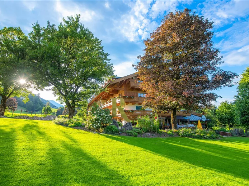 Mehrstöckiges Holzhotel mit Balkonen hinter einer grünen Wiese und großen Laubbäumen bei Sonnenschein.