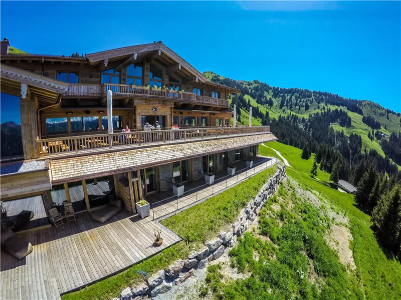 Ein großes Holzhaus in malerischer Berglandschaft. Die Sonne scheint auf eine grüne Wiese und die gegenüberliegenden Hügel.
