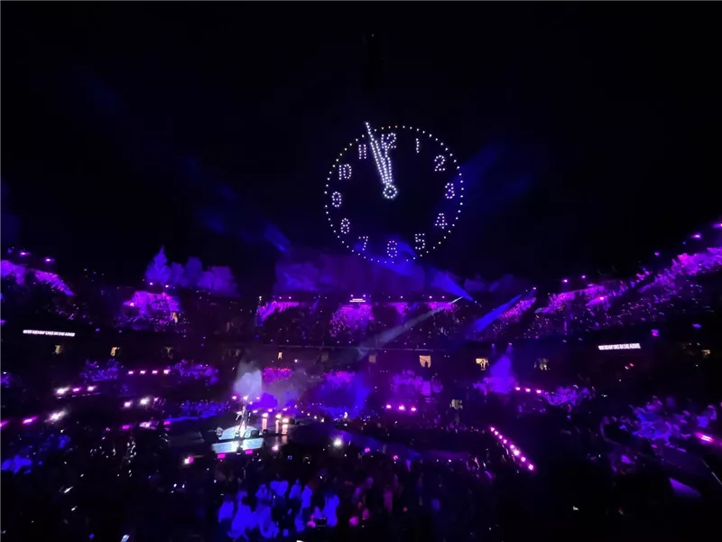 Ein aufregendes Konzert mit einer großen Menschenmenge in einer Arena. Über der Bühne hängt eine große Uhr und die Beleuchtung ist in lila Farben gehalten.