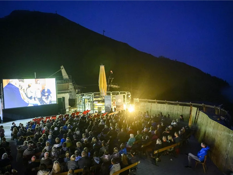Ein freiluft Kino mit einer großen Leinwand und vielen Zuschauern unter einem klaren Nachthimmel. Die Atmosphäre ist lebhaft und entspannt.