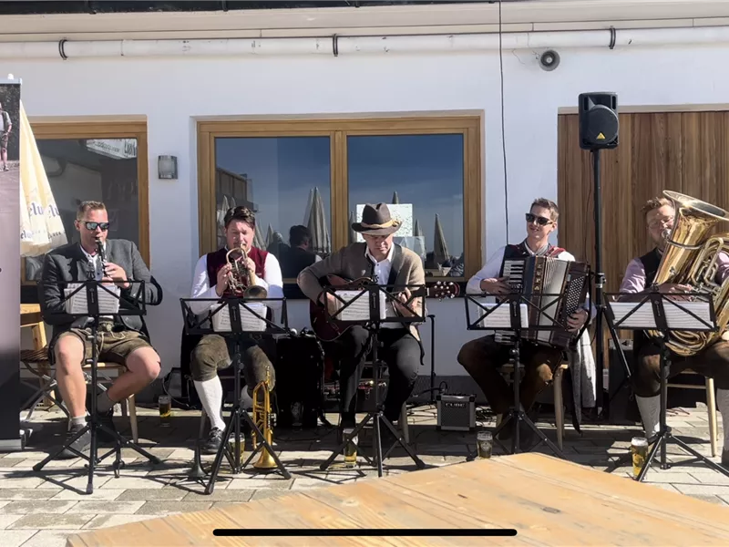 Eine Gruppe von fünf Musikern spielt traditionelle Musik im Freien. Sie tragen Trachten und umgeben sich mit Instrumenten wie Akkordeon und Posaune.