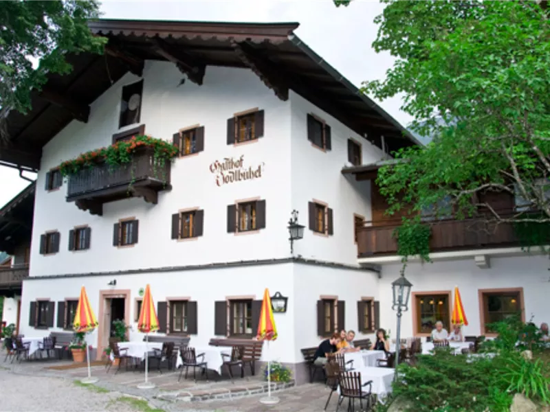 Ein traditionelles bayerisches Gasthaus mit weißer Fassade und Holzelementen. Tische im Freien mit Sonnenschirmen laden zum Verweilen ein.