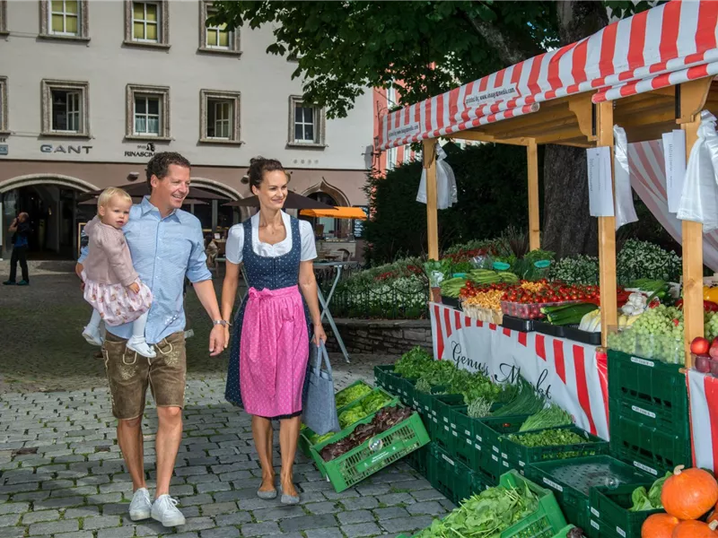 Eine Familie mit einem kleinen Kind besucht einen Bauernmarkt. Frisches Obst und Gemüse sind auf den Verkaufsständen angeboten.