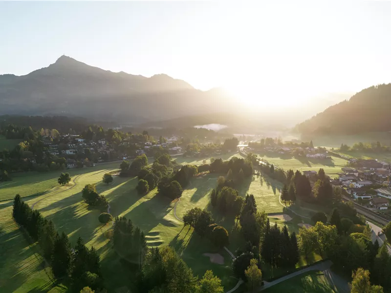 Eine malerische Golfanlage mit sanften Hügeln, Bäumen und dem Hintergrund von Bergen. Das Licht der Morgensonne lässt die Szene freundlich und einladend erscheinen.