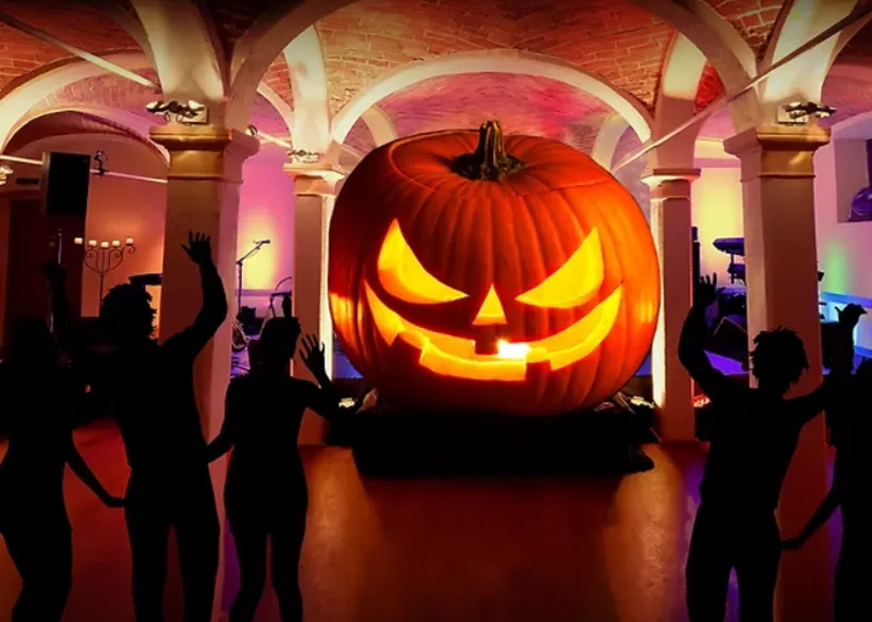 Silhouetten tanzender Personen vor einem großen, beleuchteten Halloween-Kürbis in einem Gewölbekeller.