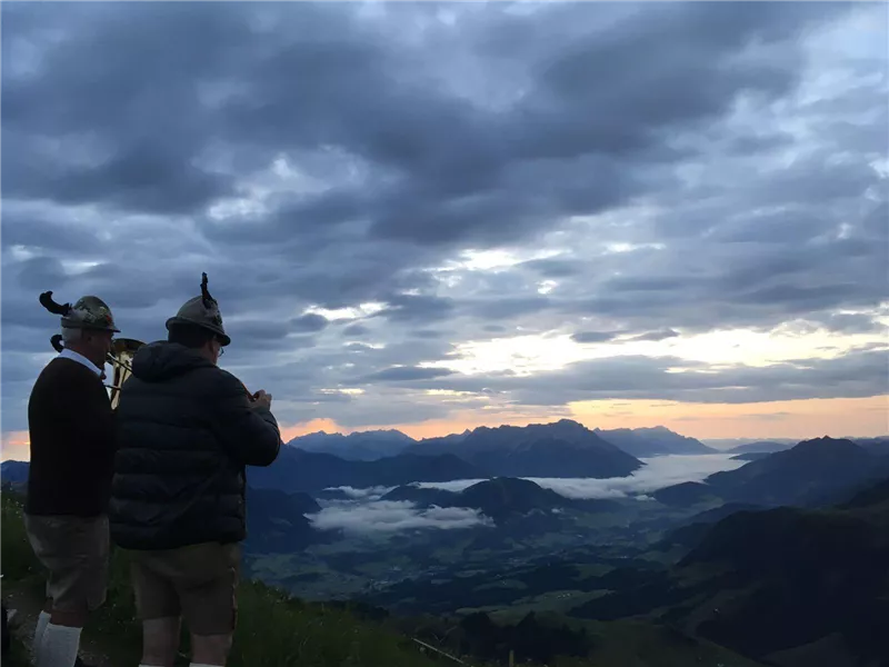 Zwei Männer in traditioneller Kleidung stehen auf einem Berg und betrachten die Landschaft. Der Himmel ist bewölkt und das Licht der Dämmerung strahlt über die Berge.