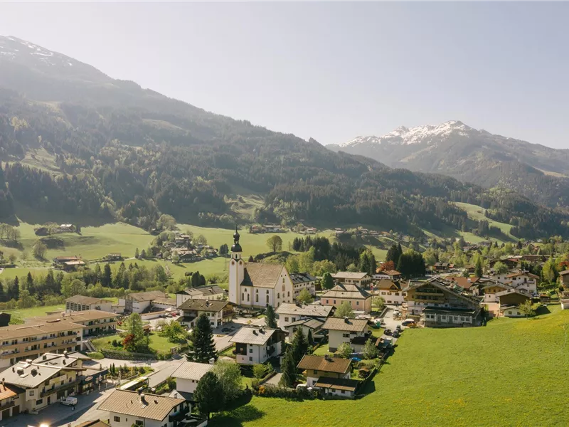 Eine malerische Berglandschaft mit einem kleinen Dorf und grünen Wiesen. Im Hintergrund sind schneebedeckte Gipfel zu sehen.