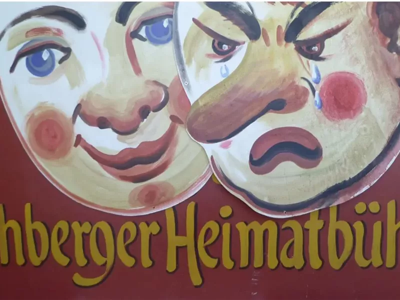Ein buntes Schild mit zwei Gesichtern, einem lächelnden und einem mürrischen Gesicht. Darunter steht der Schriftzug "Jochberger Heimatbühne".