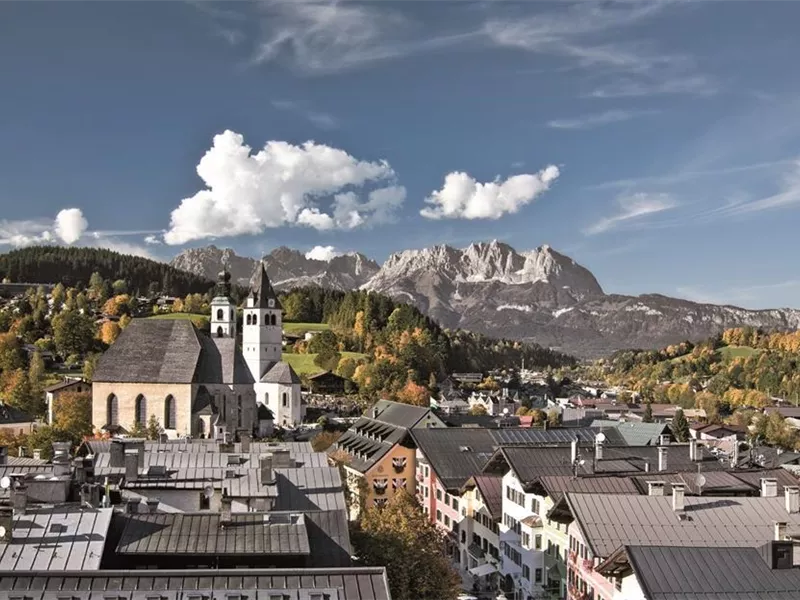 Eine malerische Stadt mit traditionellen Häusern und einer Kirche im Vordergrund. Im Hintergrund sind majestätische Berge und eine klare, blaue Himmelslandschaft zu sehen.