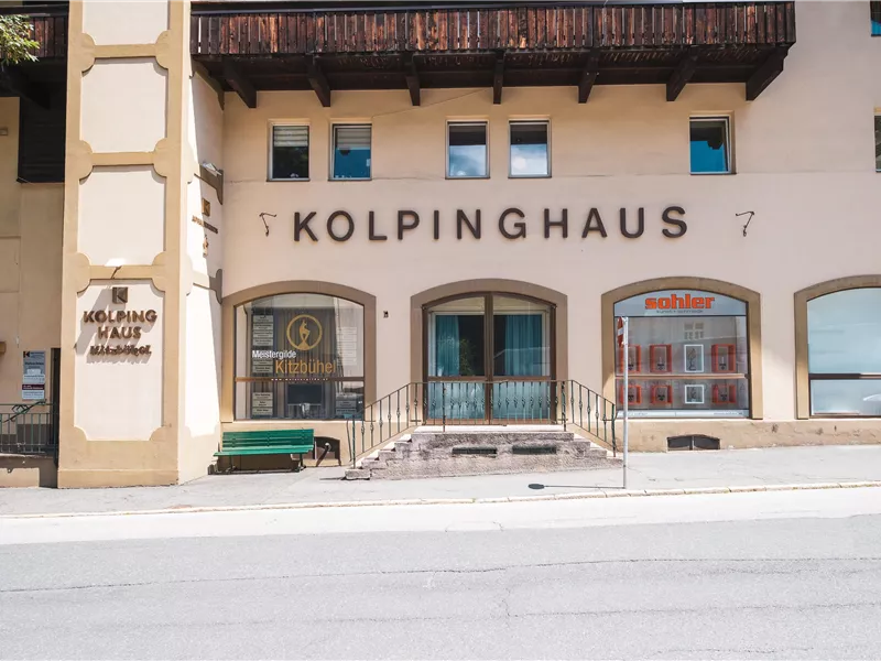 Außenansicht des Kolpinghauses in Kitzbühel mit Eingangsbereich, Schaufenstern und einer grünen Sitzbank.