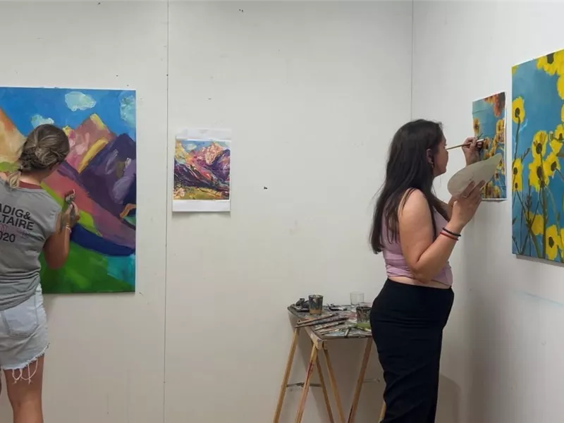 Zwei Künstlerinnen arbeiten an ihren Gemälden in einem hellen Atelier. Im Hintergrund sind bunte Landschafts- und Blumenmotive zu sehen.