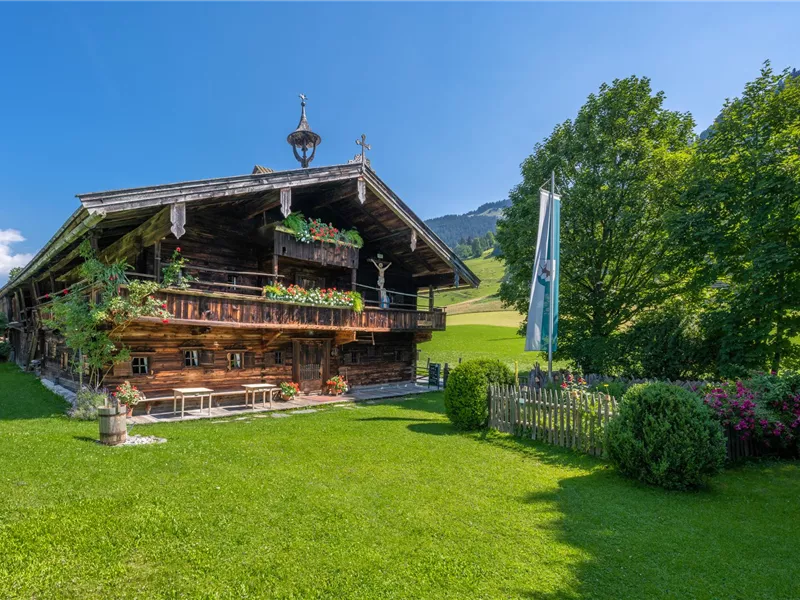 Ein gemütliches Holzhaus mit einer schönen Veranda in einer grünen Wiese. Umgeben von Bäumen und einem klaren, blauen Himmel.