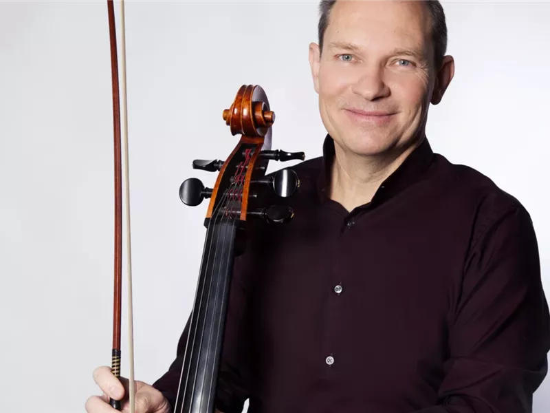 Ein Mann im dunklen Hemd hält ein Cello und einen Bogen vor weißem Hintergrund.