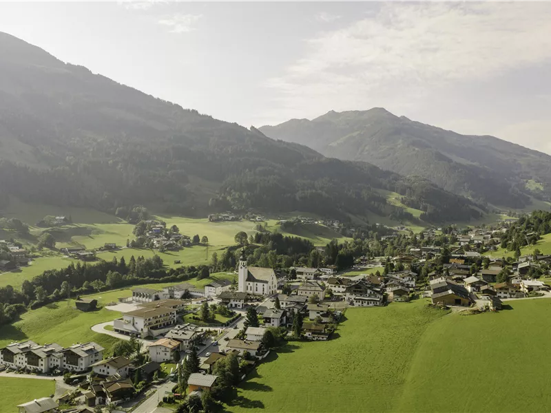 Eine malerische Landschaft mit einem Dorf im Tal und beeindruckenden Bergen im Hintergrund. Die Wiesen sind grün und die Häuser sind verstreut angeordnet.