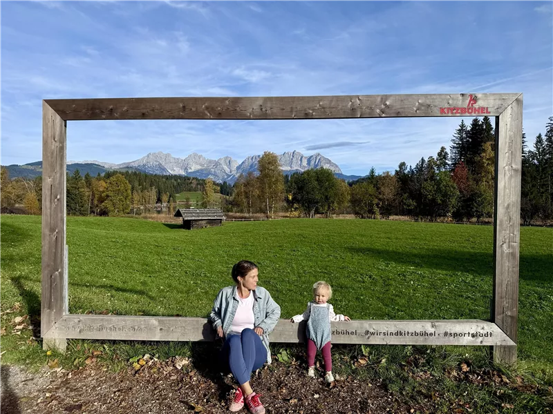 Frau und Kleinkind sitzen in einem großen Holzrahmen vor einer Bergkulisse mit Wiesen und Wald.