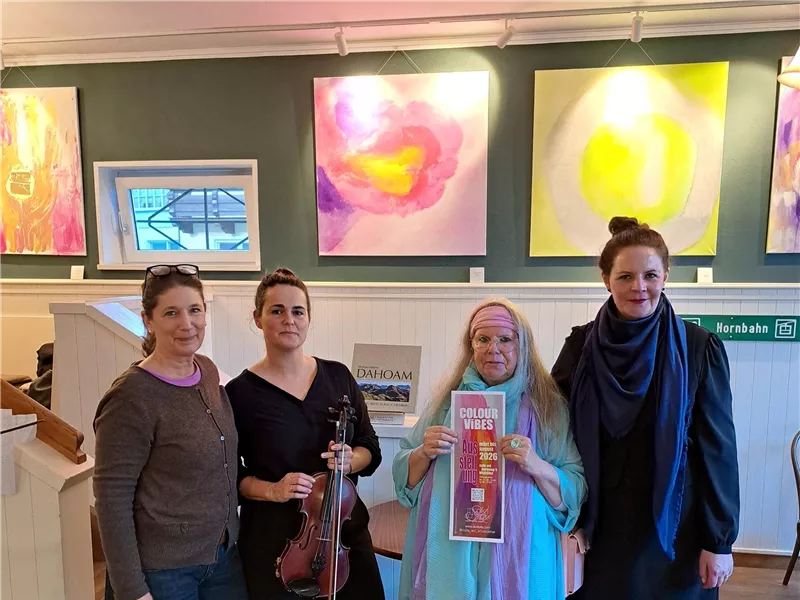 Vier Frauen stehen in einer Galerie vor bunten Gemälden. Eine Frau hält eine Geige und eine andere ein Plakat.
