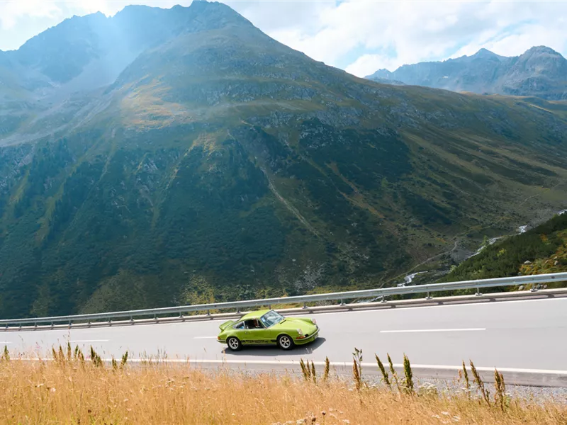 Ein grüner Sportwagen fährt auf einer asphaltierten Passstraße vor einer alpinen Bergkulisse.