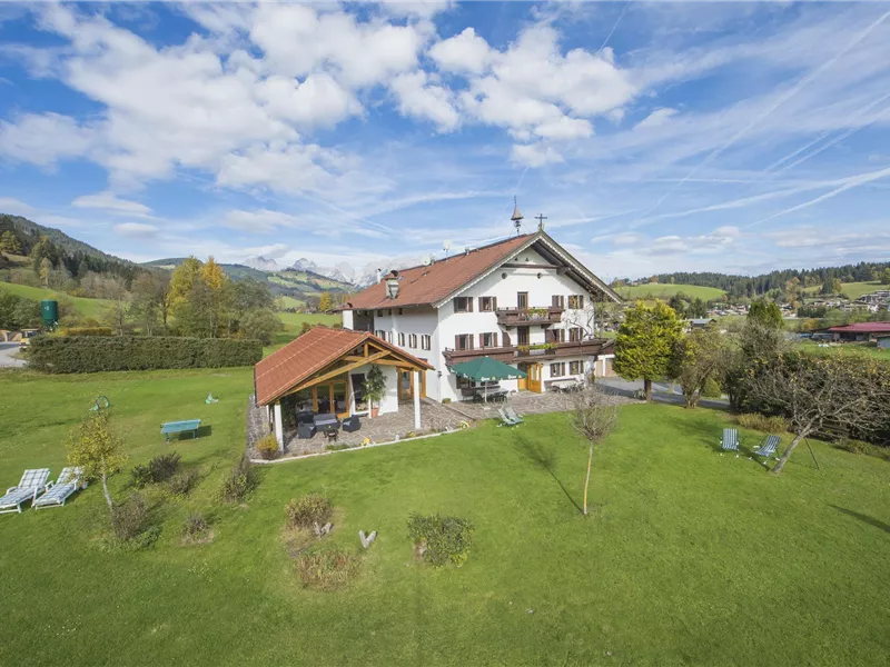 Ein malerisches Gasthaus umgeben von grüner Wiese und Bergen. Der Himmel ist blau mit einigen Wolken.