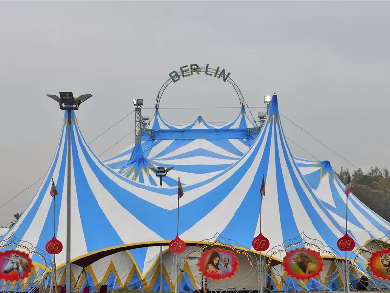 Blau-weiß gestreiftes Zirkuszelt mit dem Schriftzug Berlin auf einem Metallbogen vor grauem Himmel.