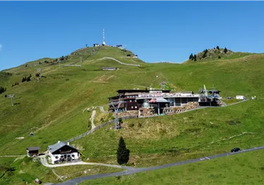 Eine grüne Berglandschaft mit einer funkelnenden Antenne auf dem Gipfel. Im Vordergrund sieht man einige Gebäude und Seilbahnen.