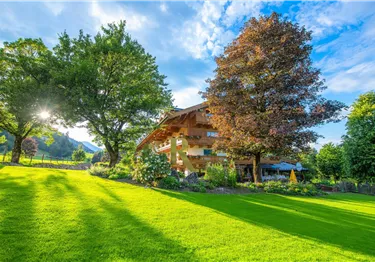 Mehrstöckiges Holzhotel mit Balkonen hinter einer grünen Wiese und großen Laubbäumen bei Sonnenschein.