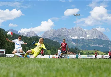 Ein Fußballspiel auf einem grünen Feld mit einem klaren Himmel und Bergen im Hintergrund. Zwei Spieler sind im Einsatz, während die Zuschauer zuschauen.