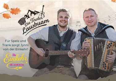 Zwei Musiker in Tracht mit Gitarre und Harmonika vor einem Holzhaus.