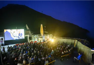 Ein freiluft Kino mit einer großen Leinwand und vielen Zuschauern unter einem klaren Nachthimmel. Die Atmosphäre ist lebhaft und entspannt.