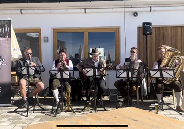 Eine Gruppe von fünf Musikern spielt traditionelle Musik im Freien. Sie tragen Trachten und umgeben sich mit Instrumenten wie Akkordeon und Posaune.