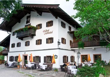 Ein traditionelles bayerisches Gasthaus mit weißer Fassade und Holzelementen. Tische im Freien mit Sonnenschirmen laden zum Verweilen ein.