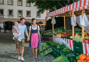 Eine Familie mit einem kleinen Kind besucht einen Bauernmarkt. Frisches Obst und Gemüse sind auf den Verkaufsständen angeboten.