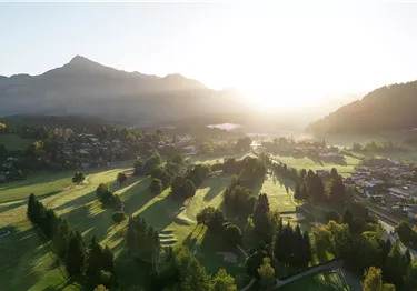 Eine malerische Golfanlage mit sanften Hügeln, Bäumen und dem Hintergrund von Bergen. Das Licht der Morgensonne lässt die Szene freundlich und einladend erscheinen.