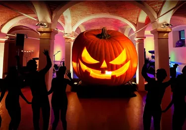 Silhouetten tanzender Personen vor einem großen, beleuchteten Halloween-Kürbis in einem Gewölbekeller.