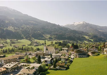 Eine malerische Berglandschaft mit einem kleinen Dorf und grünen Wiesen. Im Hintergrund sind schneebedeckte Gipfel zu sehen.