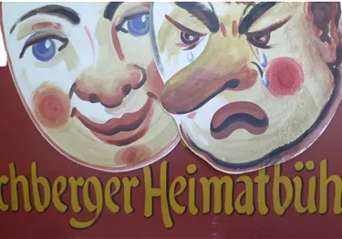 Ein buntes Schild mit zwei Gesichtern, einem lächelnden und einem mürrischen Gesicht. Darunter steht der Schriftzug "Jochberger Heimatbühne".