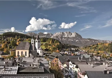Eine malerische Stadt mit traditionellen Häusern und einer Kirche im Vordergrund. Im Hintergrund sind majestätische Berge und eine klare, blaue Himmelslandschaft zu sehen.