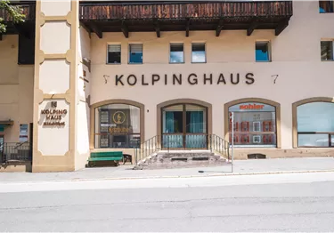 Außenansicht des Kolpinghauses in Kitzbühel mit Eingangsbereich, Schaufenstern und einer grünen Sitzbank.