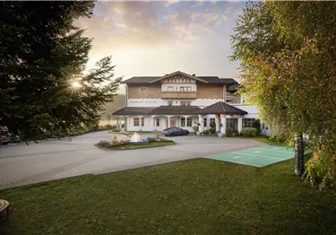 Außenansicht des Lisi Family Hotels mit gepflastertem Vorplatz, Springbrunnen und Bäumen bei Sonnenuntergang.