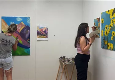 Zwei Künstlerinnen arbeiten an ihren Gemälden in einem hellen Atelier. Im Hintergrund sind bunte Landschafts- und Blumenmotive zu sehen.