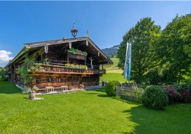 Ein gemütliches Holzhaus mit einer schönen Veranda in einer grünen Wiese. Umgeben von Bäumen und einem klaren, blauen Himmel.