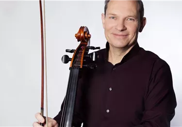 Ein Mann im dunklen Hemd hält ein Cello und einen Bogen vor weißem Hintergrund.