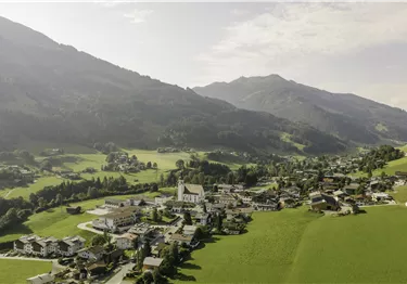 Eine malerische Landschaft mit einem Dorf im Tal und beeindruckenden Bergen im Hintergrund. Die Wiesen sind grün und die Häuser sind verstreut angeordnet.