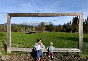 Frau und Kleinkind sitzen in einem großen Holzrahmen vor einer Bergkulisse mit Wiesen und Wald.