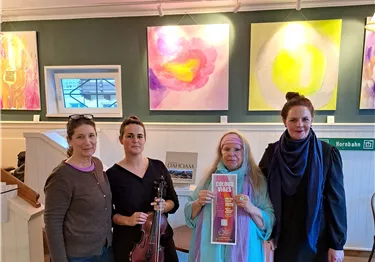 Vier Frauen stehen in einer Galerie vor bunten Gemälden. Eine Frau hält eine Geige und eine andere ein Plakat.