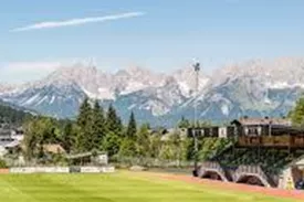 Ein Fußballplatz mit grünem Rasen und einem Sportgebäude im Hintergrund. Die majestätischen Berge sind klar am Horizont zu sehen.