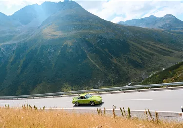 Ein grüner Sportwagen fährt auf einer asphaltierten Passstraße vor einer alpinen Bergkulisse.
