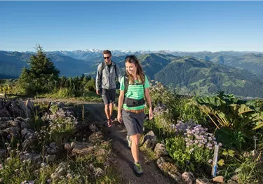 <p>Zwei Wanderer gehen auf einem Pfad durch eine bergige Landschaft mit üppiger Vegetation. Im Hintergrund sind hohe Berge und ein klarer Himmel zu sehen.</p>