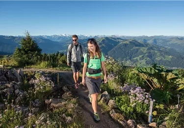 Zwei Personen wandern auf einem schmalen Bergpfad vor einem Panorama mit schneebedeckten Gipfeln.