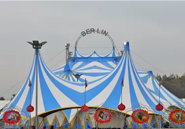 Blau-weiß gestreiftes Zirkuszelt mit dem Schriftzug Berlin auf einem Metallbogen vor grauem Himmel.
