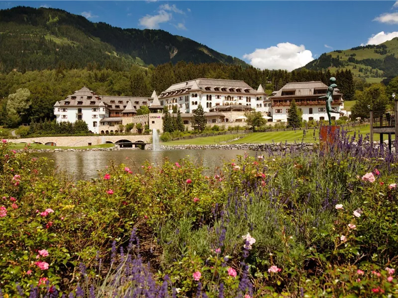 Ein malerisches Hotel am Ufer eines Sees, umgeben von blühenden Gärten. Im Hintergrund erheben sich grüne Berge unter einem klaren blauen Himmel.